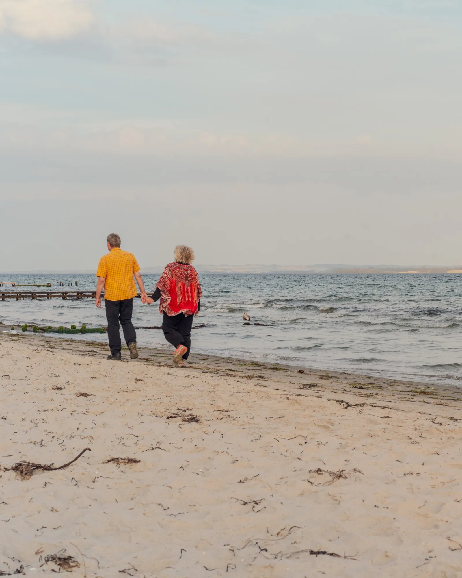 A couple walk hand in hand along an empty sandy beach toward a calm sea at golden hour.