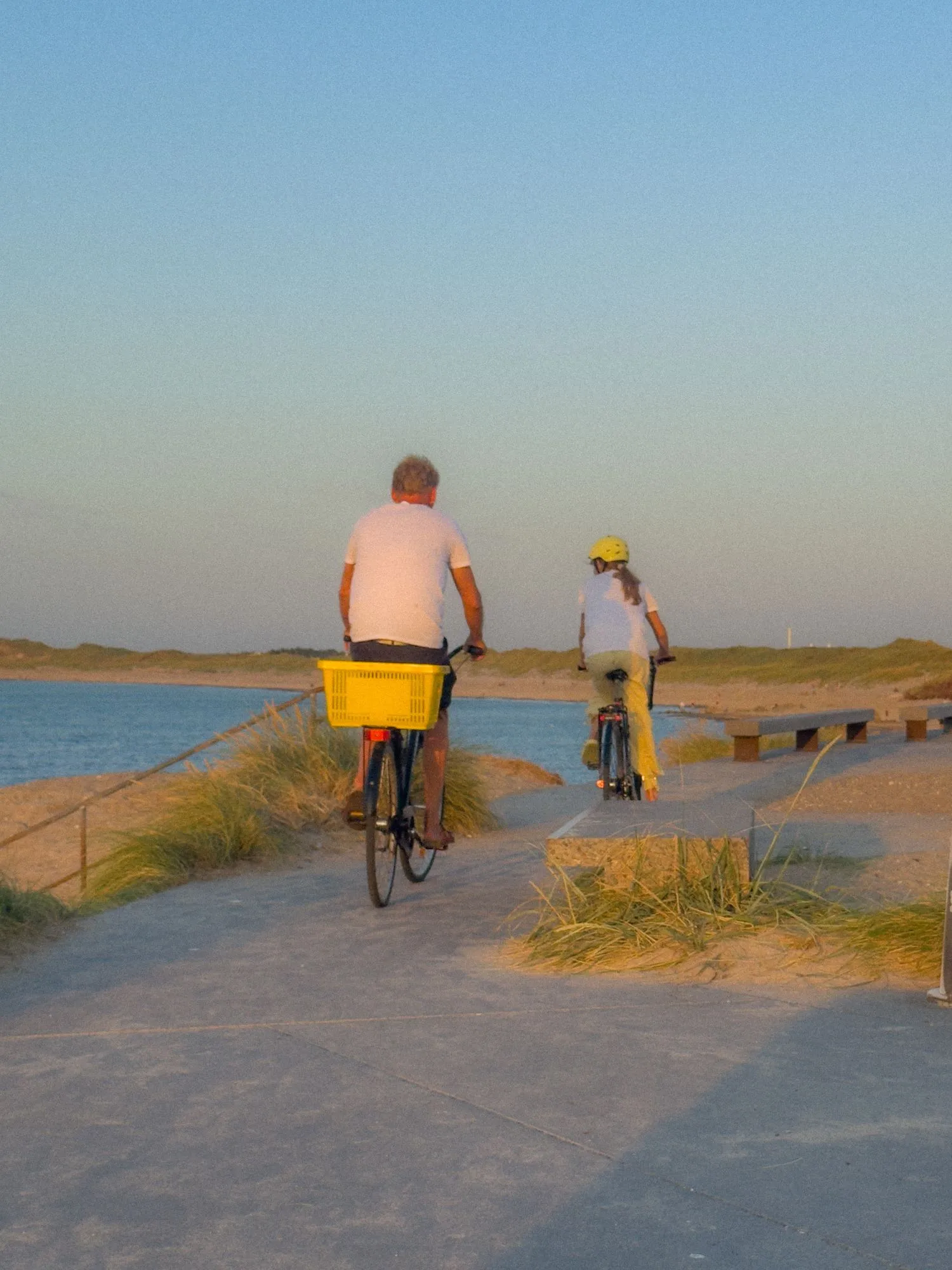 Two people cycle away down a paved path between dunes toward the sea at sunset — one with a yellow basket, the other a child in a yellow helmet.