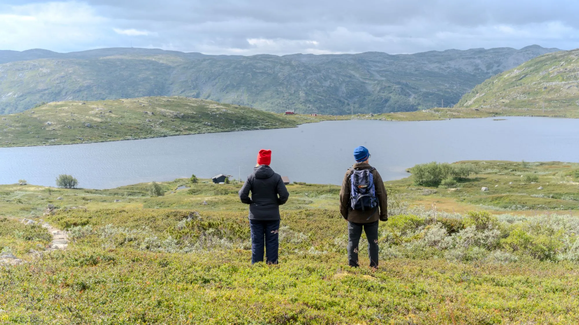 Two people stand with their backs to the camera on open moorland, looking out over a mountain lake under a grey sky.