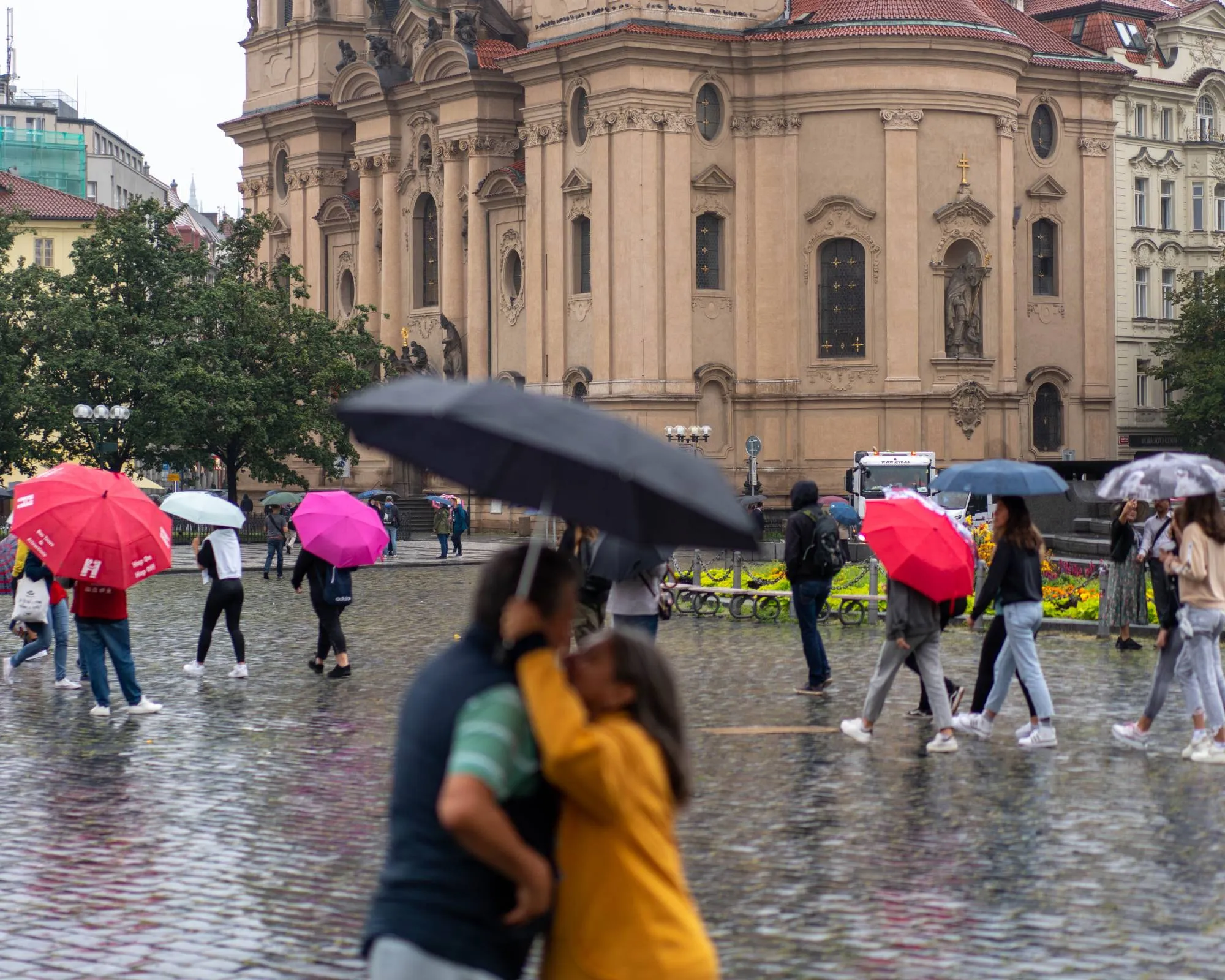 A couple embrace under an umbrella in a rainy city square while others pass with colourful umbrellas, a Baroque church behind.