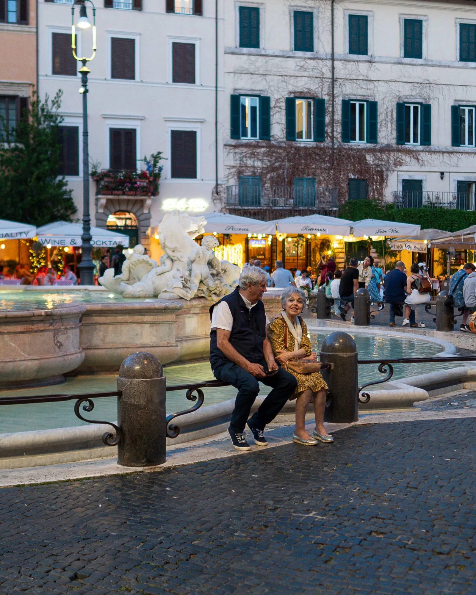 An older couple sit close together on the rim of a Baroque fountain in a Roman piazza at dusk, lit restaurants glowing behind them.